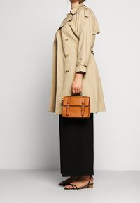 Trench-coat beige à double boutonnage avec des boutons dorés, jupe maxi noire, sandales noires à lanières, et un petit sac à main marron tenu dans la main.