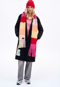 Žena, která má na sobě červenou čepici, dlouhý černý kabát s nášivkami, pruhovaný růžovo-oranžový šál, růžový svetr, šedé džíny a stříbrné tenisky.