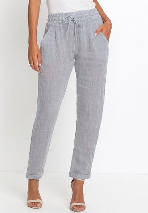 Donna che indossa pantaloni grigi e bianchi a righe verticali con coulisse e orli risvoltati, insieme a sandali beige open-toe, in piedi contro uno sfondo bianco.
