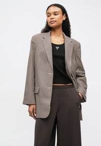 Femme portant un blazer à carreaux marron surdimensionné, un top sans manches noir avec un logo triangulaire, et un pantalon large marron foncé, se tenant avec une main dans la poche.