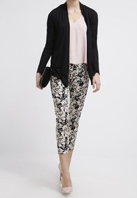 Schwarze Langarm-Cardigan über einem hellrosa Top, kombiniert mit Blumenmuster-Hosen und nudefarbenen High-Heels. Schwarze Clutch in der Hand.
