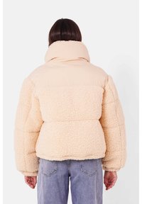 Beige shearling jakke med høj krave, pufferærmer og en glat overdel i en lysere nuance. Tekstureret, hyggeligt materiale.