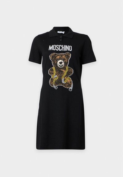 Robe polo noire avec un motif d'ours en peluche enveloppé dans un ruban à mesurer jaune et le mot MOSCHINO au-dessus de l'ours.