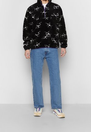 Personne portant un pull polaire noir avec des motifs d'éclairs blancs, un jean bleu et des baskets blanches avec des accents violets et beiges.