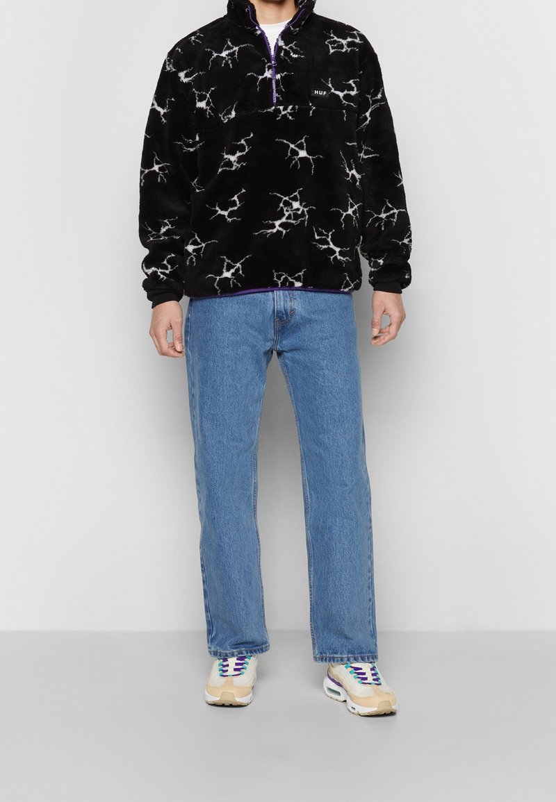 Personne portant un pull polaire noir avec des motifs d'éclairs blancs, un jean bleu et des baskets blanches avec des accents violets et beiges.