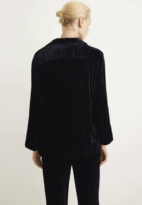 Chaqueta de terciopelo negro con un corte relajado, mangas largas y cuello. Presenta una sutil textura de canalé vertical. Sin cierre visible.