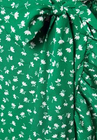 Tissu vert avec un motif floral de fleurs blanches. Présente des plis et un détail de nœud, mettant en valeur une texture douce et un matériau léger.