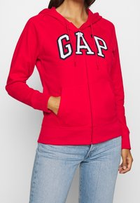 Sudadera con cremallera roja, hecha de algodón suave, que presenta un gran logo blanco de "GAP", bolsillos delanteros y capucha con cordón. Puños y dobladillo de punto acanalado básicos.