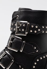 Stivale alla caviglia in pelle nera con molteplici fibbie argento e borchie, caratterizzato da un materiale testurizzato e una forma strutturata.