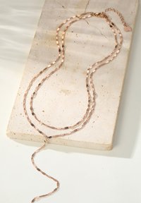 Schichtkette mit verlängerten, rechteckigen Gliedern aus Roségold mit strukturierter Oberfläche, die elegant über einer neutralen Steinoberfläche drapiert ist.