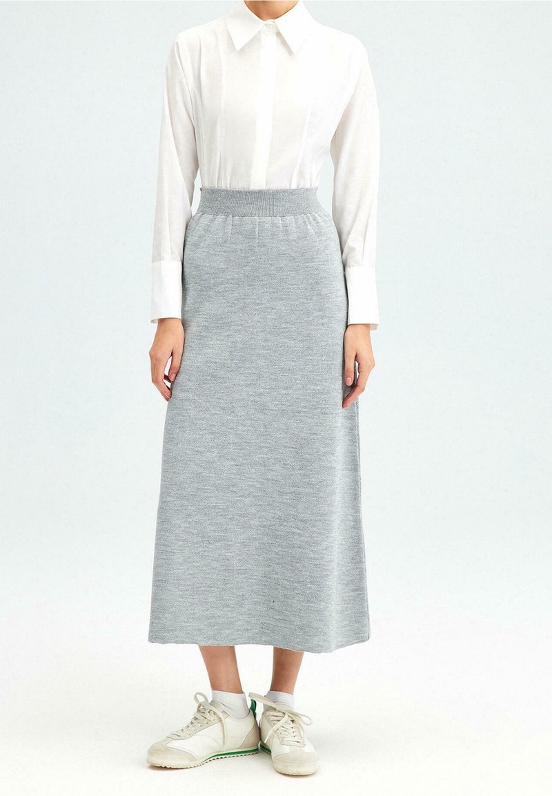 Femme portant une chemise blanche à manches longues avec col, une jupe midi en maille grise, des chaussettes blanches et des baskets blanches à semelles vertes.