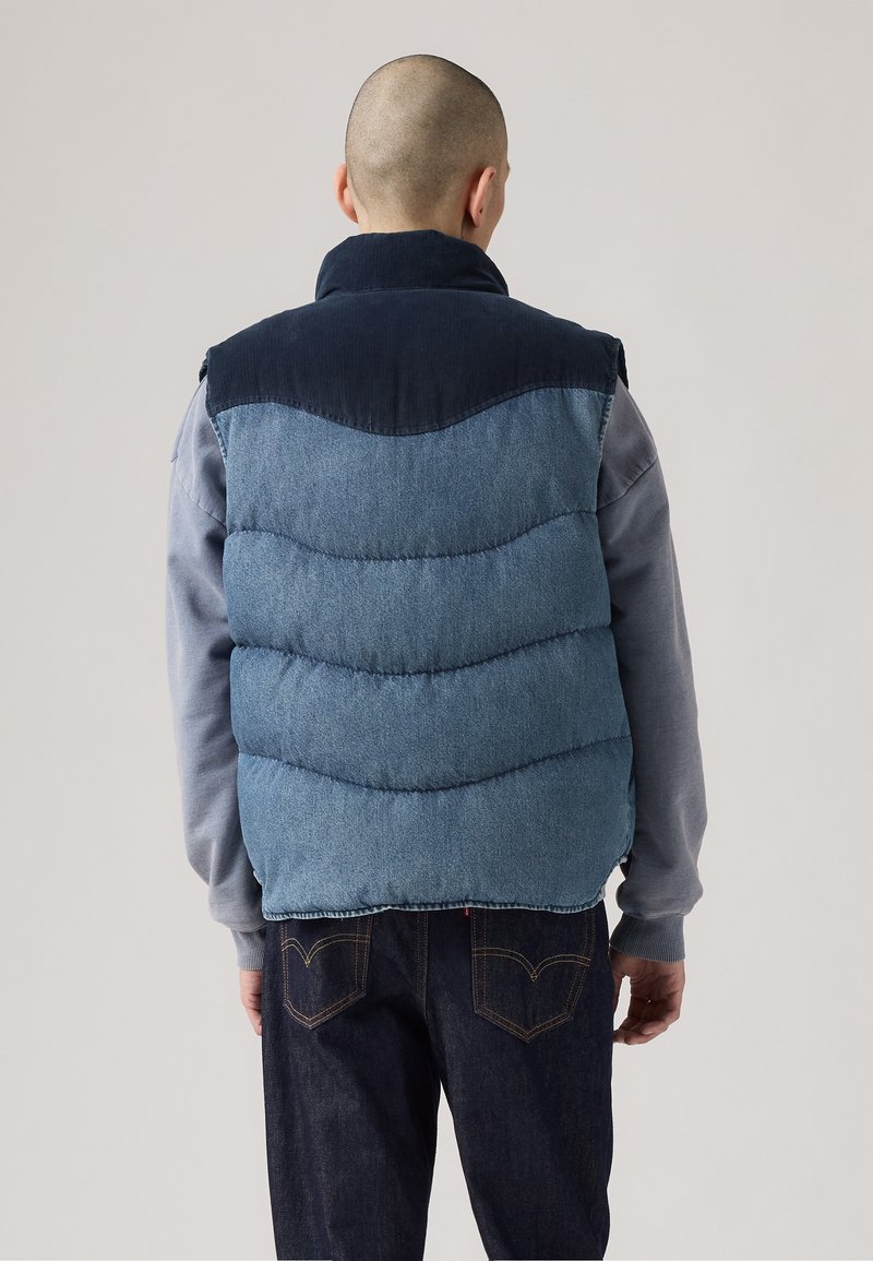 Blå denim puffer vest med en mørkeblå kordfløjlskrag, quilted design og en kant i en kontrasterende farve. Bagfra ses syning detaljerne.