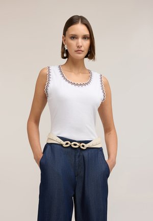 Femme aux cheveux courts bruns portant un haut blanc sans manches avec des bords festonnés, un pantalon bleu foncé, une ceinture beige et des boucles d'oreilles en perles, les mains dans les poches.