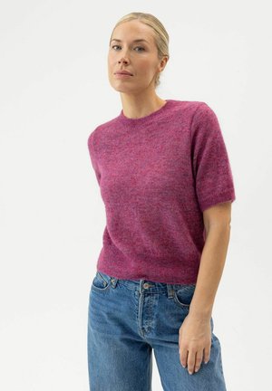 Kvinde med blondt hår iført en lilla kortærmet sweater og blå jeans, stående mod en hvid baggrund.
