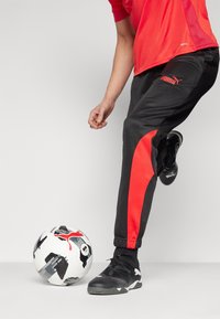 Melni un sarkani sporta bikses ar sašaurām kājām un elastīgām aprocēm, kopā ar melniem sporta apaviem, blakus melnbaltai rakstainai futbola bumbai.