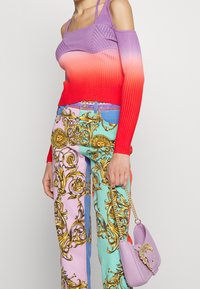Gerippter Pullover in lila-rotem Farbverlauf mit ausgeschnittenen Schultern; gemusterte Hose in Pastellfarben mit goldenen barocken Designs; kleine lavendelfarbene Handtasche.