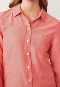 Chemise corail boutonnée en tissu léger. Elle présente un col, des manches longues et une poche poitrine unique. Les boutons blancs sont visibles.