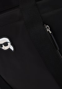 Zwarte tas gemaakt van gladde stof, met een rits en een klein wit grafisch label met een karakter in een zonnebril.