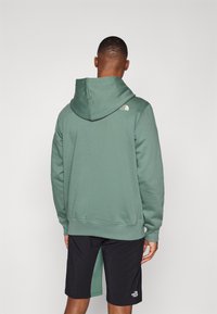 Hanorac cu glugă de culoare verde salvie, realizat dintr-un material moale, având un logo pe umărul stâng. Asortat cu pantaloni scurți negri.
