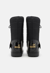 Melnas šūtās ziemas zābaki, kuriem ir spīdīgs melns ādas apakšdaļa ar zelta "LOVE MOSCHINO" zīmējumu un sānu rāvējslēdzēja detaļu.