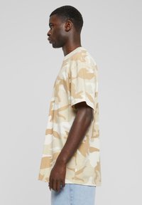Beige camouflage T-shirt med korte ærmer og en afslappet pasform, lavet af blødt stof. Funktioner inkluderer en rund halsudskæring og subtile farvevariationer.