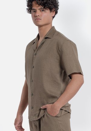 Giovane uomo con capelli ricci che indossa una camicia marrone a maniche corte con bottoni e pantaloni abbinati, in piedi con una mano nella tasca su sfondo bianco.