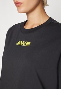 Mustmust jakk, mis on valmistatud puuvillast, ümmarguse kaelusega ja esiküljel kollane "WB" logo, sileda tekstuuri ja minimalistikuga disainiga.