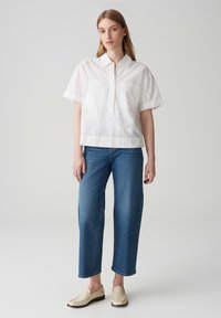 Weißes kurzärmliges Hemd mit zwei Brusttaschen, kombiniert mit blauen Jeans im geraden Schnitt und beigen Mokassins, vor einem schlichten Hintergrund.