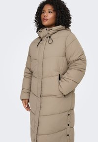 Beige vadderad jacka med huva, som har en quiltad design, dragkedjeförsedda sidofickor och tryckknappar längs fram.