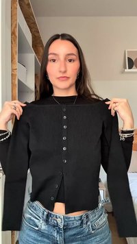 Νεαρή γυναίκα κρατά ένα μαύρο κουμπωτό ζακέτα, δείχνοντάς το από μπροστά, φορώντας μαύρο τοπ και μπλε τζιν, σε ένα δωμάτιο.