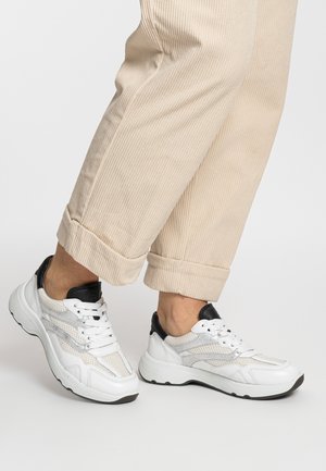 White athletic sneakers with mesh panels, black accents, and textured soles, worn with beige corduroy pants, featuring turned-up cuffs.