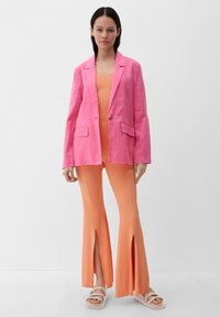Heldere roze linnen blazer met twee voorzakken, gecombineerd met een oranje geribbeld jumpsuit met gespleten broekspijpen. Lichte schoeisel maakt de look compleet.