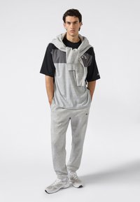 Joven vestido con pantalones deportivos grises, camiseta negra y gris, sudadera gris sobre los hombros y zapatillas blancas, de pie con las manos en los bolsillos.