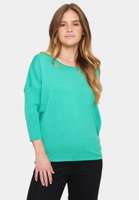 Mėtų žalios spalvos, laisvo kirpimo megztinis su trimis ketvirtadaliais rankovėmis ir apvaliu kaklu. Ribbingo tekstūra ir atsipalaidavusi siluetas. Dėvima su juodomis kelnėmis.