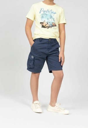 Barn står med hænderne i lommerne iført en gul T-shirt med grafik, marineblå cargoshorts og hvide sneakers på en ensfarvet baggrund.