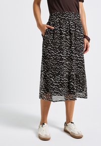 Personne portant une jupe midi noire à motifs, un haut noir, des baskets blanches, avec une main dans la poche de la jupe et des bracelets au poignet.