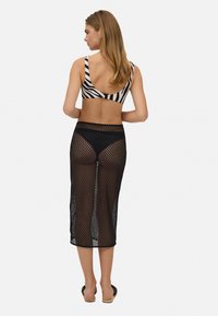 Falda de red negra con una textura transparente, combinada con un top de bikini de rayas de cebra. Se llevan pantuflas en los pies; el fondo es blanco liso.