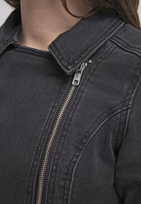 Veste en denim noir avec un col à pression et une fermeture éclair à l'avant. Le tissu présente une texture visible avec des accents de couture le long des coutures.