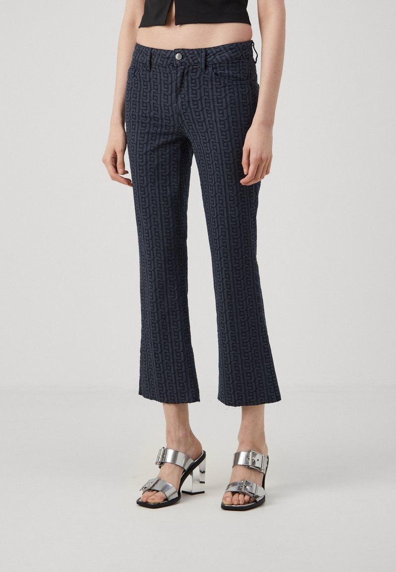 Pantalon court bleu foncé avec un motif monogramme subtil, coupe droite et ourlet effiloché. Associé à des sandales à talons hauts argentées.