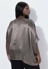 Metallisch bronzefarbenes Oversized-Oberteil mit lockerer Passform, kurzen Ärmeln, strukturiertem Stoff und abgerundetem Saum, kombiniert mit schwarzen Hosen.