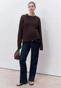 Mujer con un suéter de punto marrón oscuro, jeans azul oscuro, chanclas negras, sosteniendo un bolso marrón, de pie contra una pared blanca lisa.