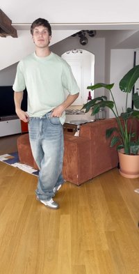 Svijetlozeleni oversize majica, plave izblijedjele traperice, srebrne tenisice. Prostorija ima teksturiranu narančastu sofu i saksiju s biljkom na drvenom podu.