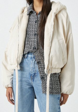 Femme portant une veste à capuche beige clair avec doublure en fourrure sur une chemise boutonnée à motifs noir et blanc et un jean bleu clair taille haute.
