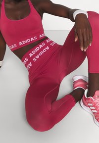 Soutien-gorge de sport rouge avec le logo "ADIDAS" en blanc, leggings taille haute assortis, et chaussures de sport roses avec des accents blancs. Tissu lisse et ajusté.