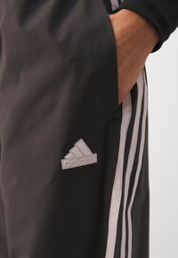 Ręka w kieszeni, ubrana w czarne spodnie dresowe Adidas z białymi paskami i logo Adidas na lewej udzie.