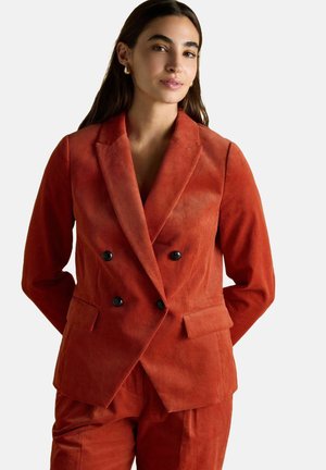 Woman with long dark hair wearing an orange corduroy double-breasted blazer with black buttons and matching pants, standing with hands behind back.
