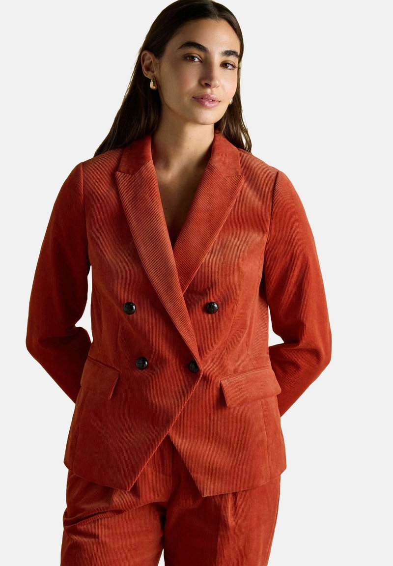 Sieviete ar gariem tumšiem matiem, tērpta oranžā korduroja divrindu žaketē ar melniem pogām un saskaņotās biksēs, stāv ar rokām aiz muguras.