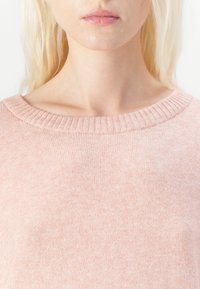 Světle růžový svetr s žebrovaným výstřihem, měkkou texturou a mírně volným střihem. Materiál se jeví jako hladký a lehký.
