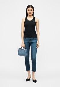 Γυναίκα με μαύρη αμάνικη μπλούζα και κοντό τζιν παντελόνι, κρατώντας μια μικρή μπλε τσάντα, στέκεται μπροστά σε ένα απλό λευκό φόντο.