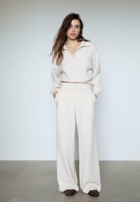 Frau steht mit Händen in den Taschen, trägt cremefarbene, weite Hosen, ein passendes Langarmoberteil mit Kragen und dunkle Schuhe vor einem schlichten Hintergrund.